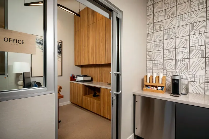 Leasing Office Entry With Glass Door, Wood Cabinetry, Coffee Station, Patterned Wall Tile, And Modern Lighting.