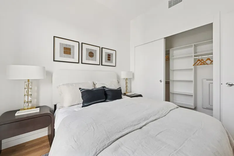 Bright Bedroom With White Bedding, Framed Wall Art, Wood Flooring, Table Lamps, And Open Closet Shelving.